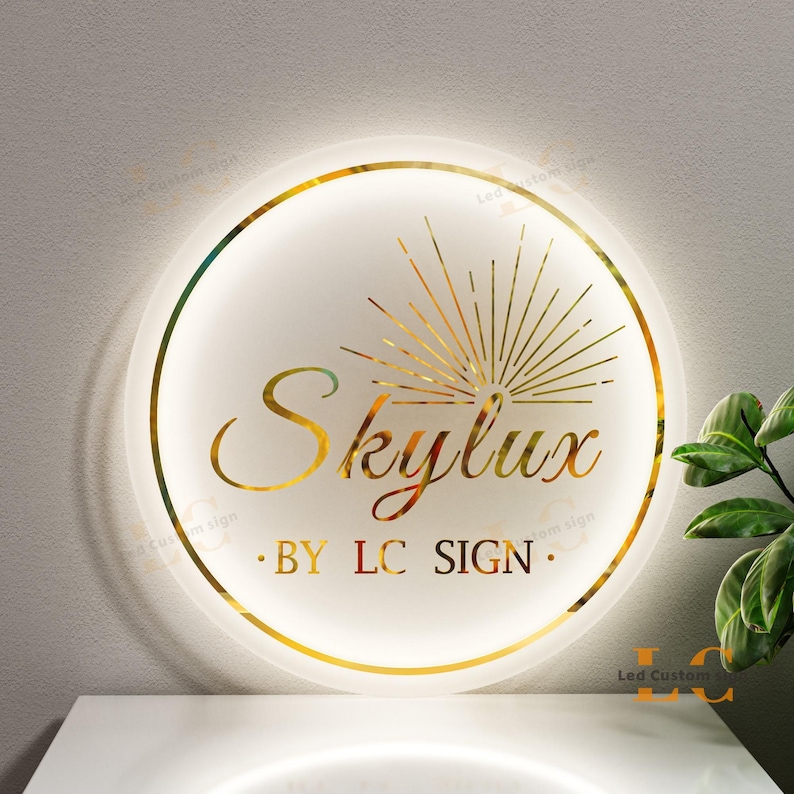 May include: A round, illuminated sign with a gold border and the name "Skylux" in elegant script. The sign also features the text "BY LC SIGN" and a gold sunburst design. The sign is mounted on a white surface.