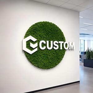 May include: A circular wall sign featuring a dense, vibrant green moss texture as a background. Mounted on the moss is a large, white, three-dimensional logo consisting of a geometric hexagon-based icon followed by the word CUSTOM in capital letters.