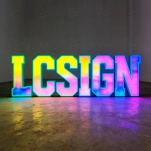 May include: Large, illuminated sign spelling out "LCSIGN". The letters are outlined in blue and filled with a gradient of colors, including yellow, pink, and blue. The sign is placed on a concrete floor.