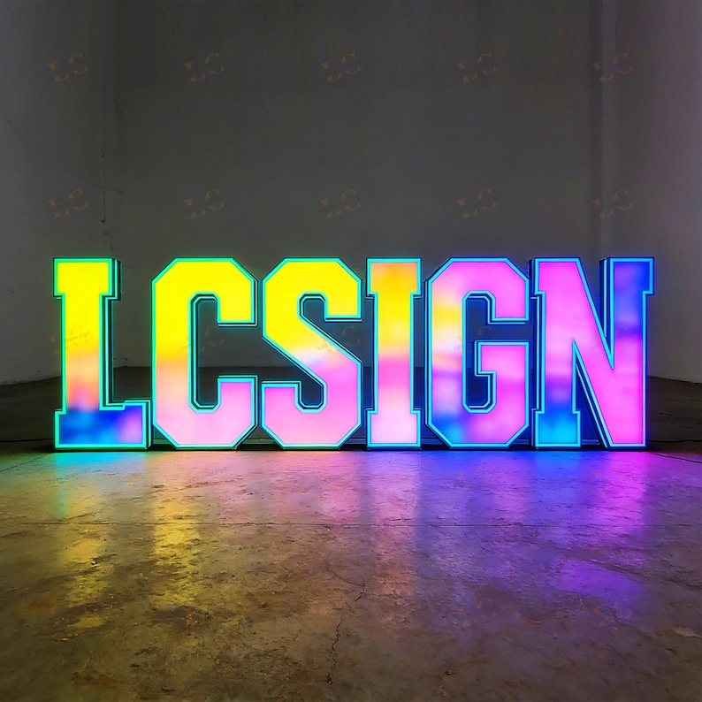 May include: Large, illuminated sign spelling out "LCSIGN". The letters are outlined in blue and filled with a gradient of colors, including yellow, pink, and blue. The sign is placed on a concrete floor.