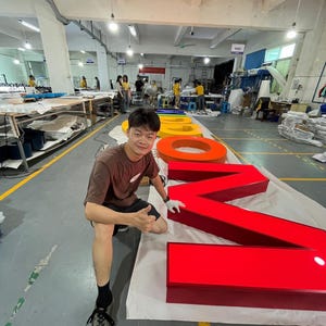 May include: Large, three-dimensional channel letters for a custom sign, featuring a bright red letter M, a red letter O, and a yellow letter O, laid out on a white protective sheet on a factory floor, with the LC Sign logo visible in the corner.
