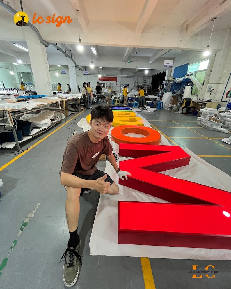 May include: Large, three-dimensional channel letters for a custom sign, featuring a bright red letter M, a red letter O, and a yellow letter O, laid out on a white protective sheet on a factory floor, with the LC Sign logo visible in the corner.