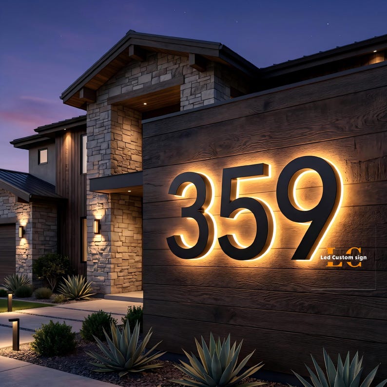 May include: Illuminated black house numbers 359 mounted on a wooden wall. The numbers are backlit with a warm, inviting glow. The house exterior features stone and wood siding, with landscaping and exterior lighting.
