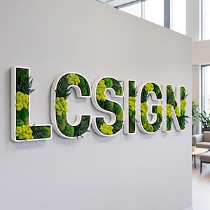 May include: A wall-mounted sign spelling the word LCSIGN in large white capital letters. Each letter is filled with preserved green moss and dark green fern leaves, creating a textured, natural appearance against the light gray wall surface.
