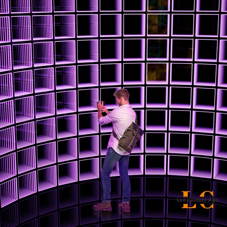 May include: A person wearing a white button-down shirt, blue jeans, and a brown backpack stands before a large, curved wall installation composed of a grid of square frames illuminated with bright purple LED lights, creating an infinity effect.