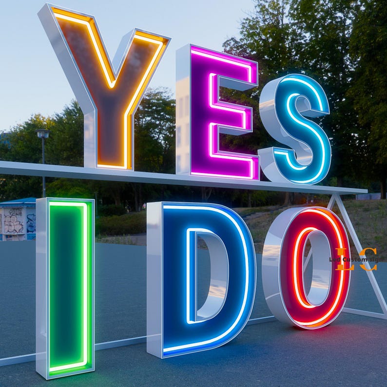 K&ouml;nnte beinhalten: Gro&szlig;es Leuchtschild mit der Aufschrift "YES I DO" mit Neonlichtern. Die Buchstaben sind wei&szlig; mit farbigen Neonkonturen: gelb, pink, blau, gr&uuml;n, blau und rot. Das Schild wird im Freien gezeigt.