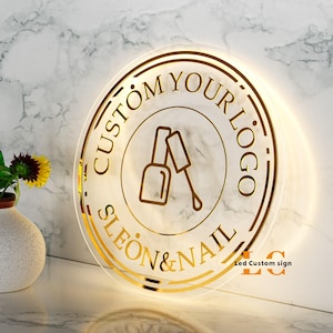 May include: A round, illuminated sign with gold lettering and a nail polish bottle and brush icon. The sign reads "CUSTOM YOUR LOGO" and "SLEON & NAIL". The sign is lit with a warm glow and has the text "LED Custom sign".