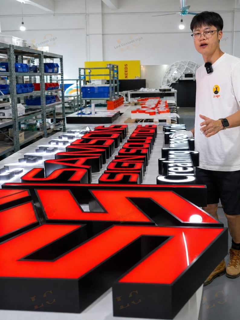 May include: A workshop scene featuring illuminated LED signs in various stages of completion. The signs are primarily red and black, with some white. The signs spell out words and phrases. A person stands next to the signs.