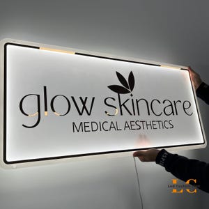 May include: A rectangular illuminated sign with the words "glow skincare" in a stylish font, along with the text "MEDICAL AESTHETICS". The sign has a black border and is being held up by two hands.