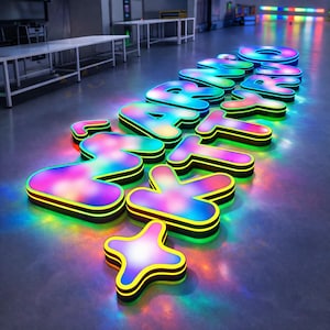 May include: Illuminated sign spelling out the word "START NOW" in large, colorful, 3D letters. Each letter has a rainbow gradient and is outlined in black and yellow. The sign is on a reflective surface.
