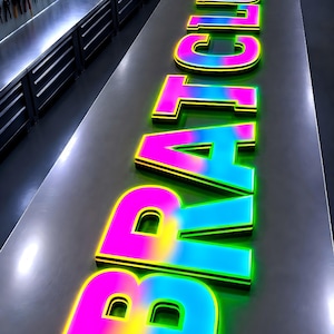 Puede incluir: Un letrero luminoso con la inscripci&oacute;n "BRAT CLUB". Las letras son grandes y multicolores, con una transici&oacute;n de rosa a amarillo a azul, delineadas en negro y verde. El letrero est&aacute; sobre una superficie met&aacute;lica.