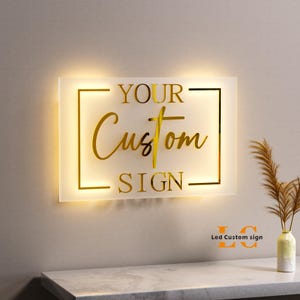 May include: A rectangular, illuminated sign with the words "YOUR Custom SIGN" in gold lettering. The sign is lit from behind, creating a warm glow. The words are inside a gold rectangular frame. The sign is mounted on a wall.