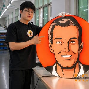 May include: A large, round custom sign featuring a stylized portrait of a man with an orange background. The sign has the text "LC Custom signs" in the bottom right corner. A person is holding the sign.