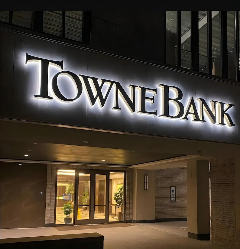 May include: A building exterior featuring a large, illuminated sign that reads TowneBank in black serif lettering with a bright white backlight. The sign is mounted above a glass-paneled entrance door, with brick pillars and interior lighting visible.