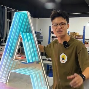 May include: A large, illuminated letter "A" with a gradient of blue and white lights. The letter is framed in a silver border. The background shows a person in a green shirt with a logo.