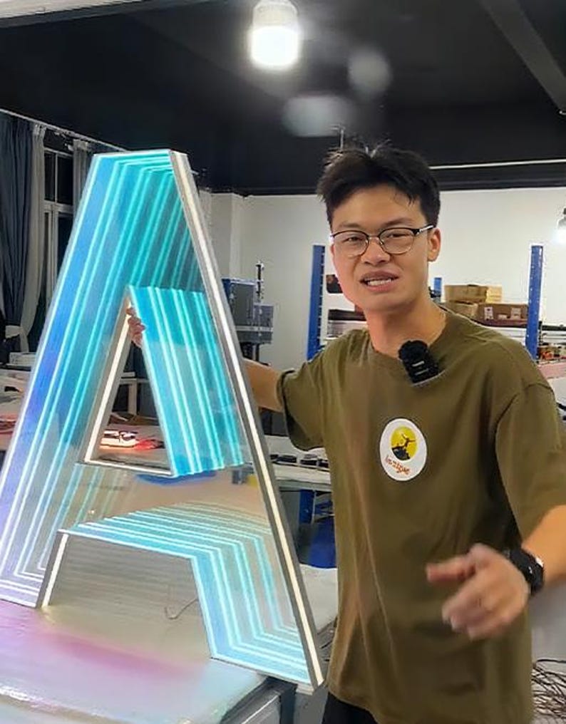 May include: A large, illuminated letter "A" with a gradient of blue and white lights. The letter is framed in a silver border. The background shows a person in a green shirt with a logo.