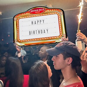 May include: A marquee sign with the words "HAPPY BIRTHDAY" in white letters. The sign has a red and gold border and is held up in a crowded room. The sign is from Buffalo Bills.