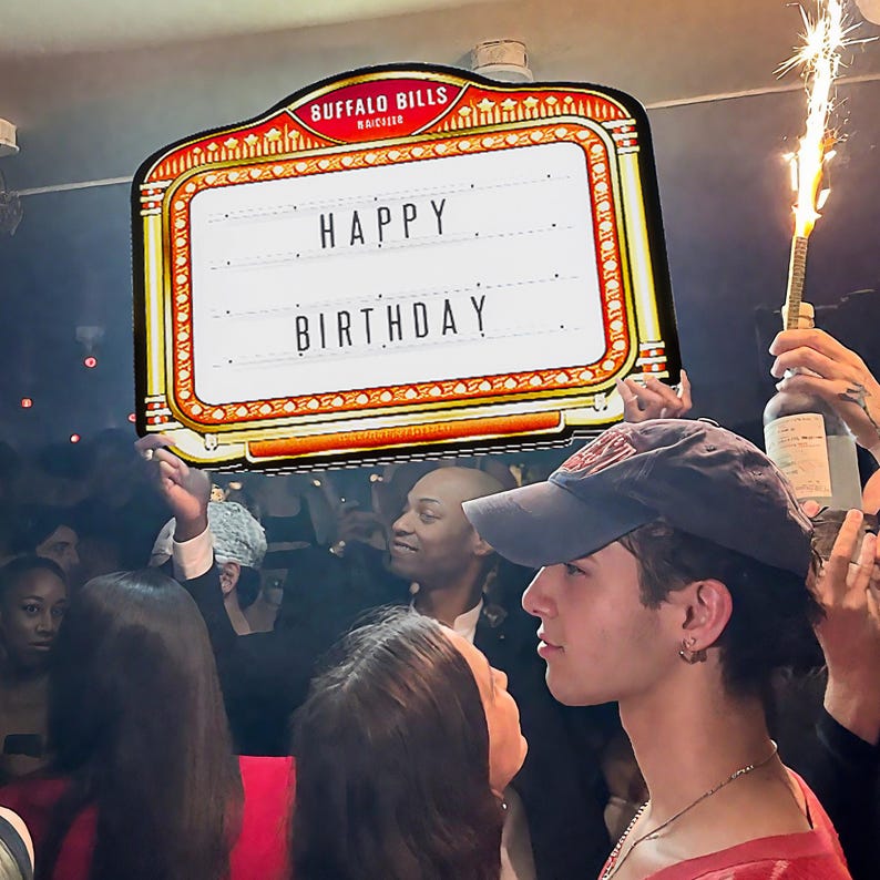 May include: A marquee sign with the words "HAPPY BIRTHDAY" in white letters. The sign has a red and gold border and is held up in a crowded room. The sign is from Buffalo Bills.