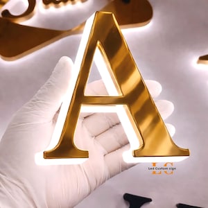 May include: A three-dimensional letter A sign with a polished gold metallic finish and integrated white LED backlighting. The letter features a clean, serif-style font and emits a soft glow from behind its edges. Text reads Led Custom sign.