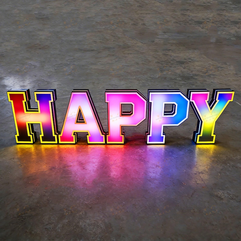 May include: A colorful, illuminated sign spells out the word "HAPPY". The letters are outlined in black and yellow, with a gradient of colors filling each letter, including red, pink, purple, blue, and yellow. The sign is on a gray surface.