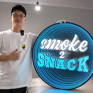 May include: A circular LED sign featuring a black frame and a blue concentric ring pattern. The text "smoke 2 SNACK" is displayed in the center in a layered, three-dimensional white and blue font, creating an optical illusion of depth.
