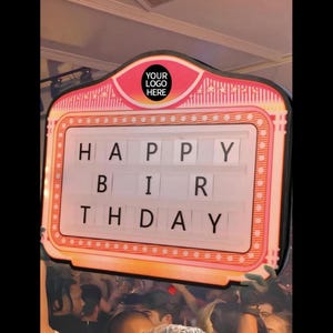 May include: A pink and white illuminated sign with the words "HAPPY BIRTHDAY" displayed in black letters. The sign has a retro design with a space for a logo at the top. The sign is held up in a crowd.