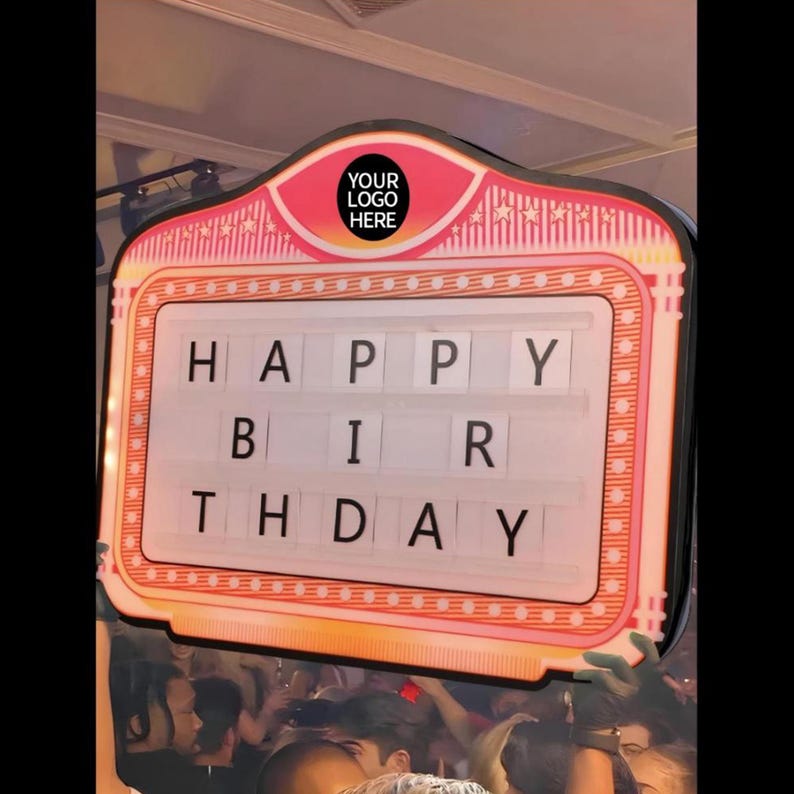 May include: A pink and white illuminated sign with the words "HAPPY BIRTHDAY" displayed in black letters. The sign has a retro design with a space for a logo at the top. The sign is held up in a crowd.