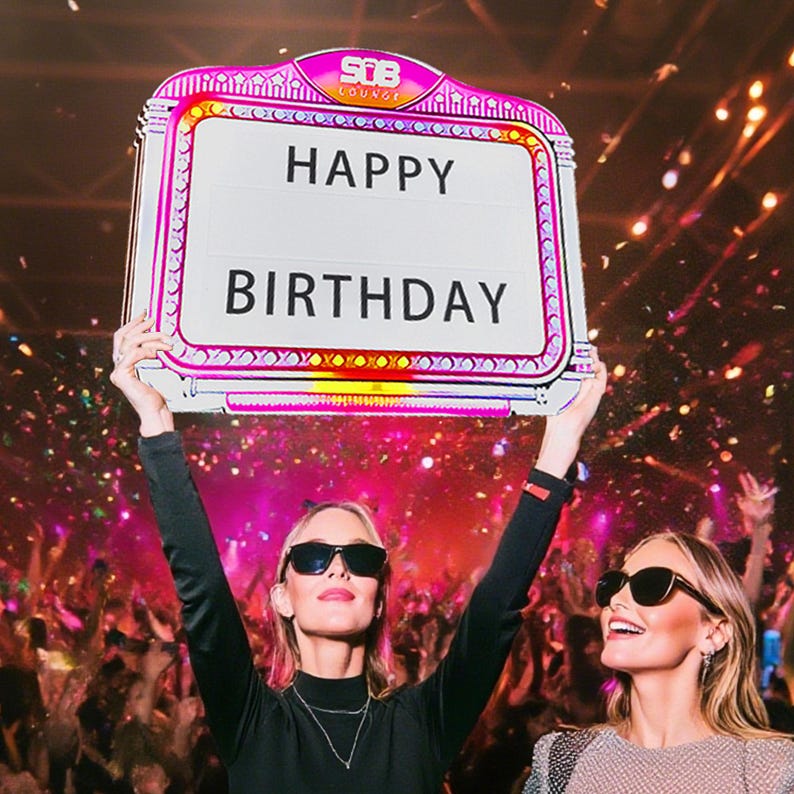 May include: A sign with the words "HAPPY BIRTHDAY" in bold, white letters. The sign has a pink and white border with decorative lights. Two people are holding the sign up in a crowded, celebratory setting.