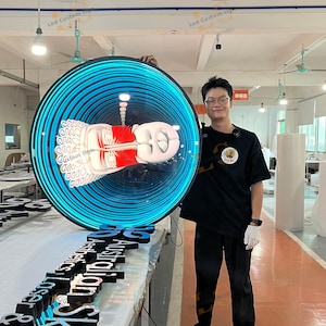 Puede incluir: Un gran letrero LED redondo con un borde de neón azul y un gráfico central de una estatua de Buda. El letrero lo sostiene una persona en un taller. Letras negras "Australian Aesthetics Lasers" sobre una mesa.