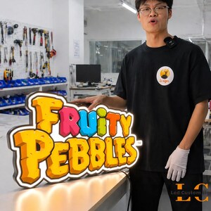 May include: A colorful LED sign featuring the text "Fruity Pebbles" in a playful font. The sign has a yellow and orange color scheme with a white border and is illuminated with warm white lights. The sign is on a table.