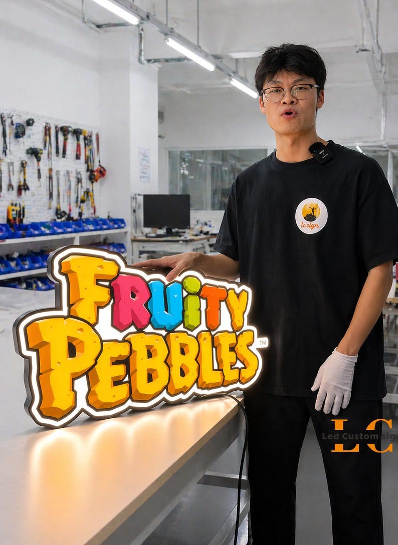 May include: A colorful LED sign featuring the text "Fruity Pebbles" in a playful font. The sign has a yellow and orange color scheme with a white border and is illuminated with warm white lights. The sign is on a table.