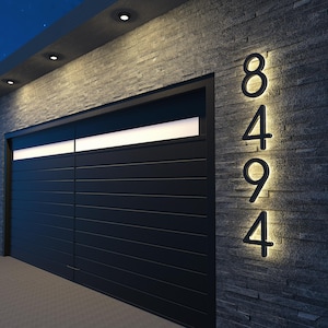 May include: Illuminated house numbers 8494 mounted on a dark gray stone wall. The numbers are black and backlit with a warm glow. A black garage door is to the left, with a horizontal white light above.