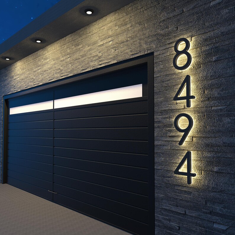 May include: Illuminated house numbers 8494 mounted on a dark gray stone wall. The numbers are black and backlit with a warm glow. A black garage door is to the left, with a horizontal white light above.