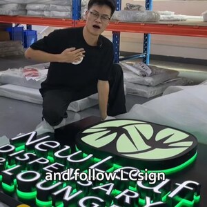 May include: A black rectangular sign featuring the white text New Leaf Dispensary Lounge and a light green leaf logo. The sign has bright green LED backlighting behind the raised letters and logo. The text and follow LCsign is visible.