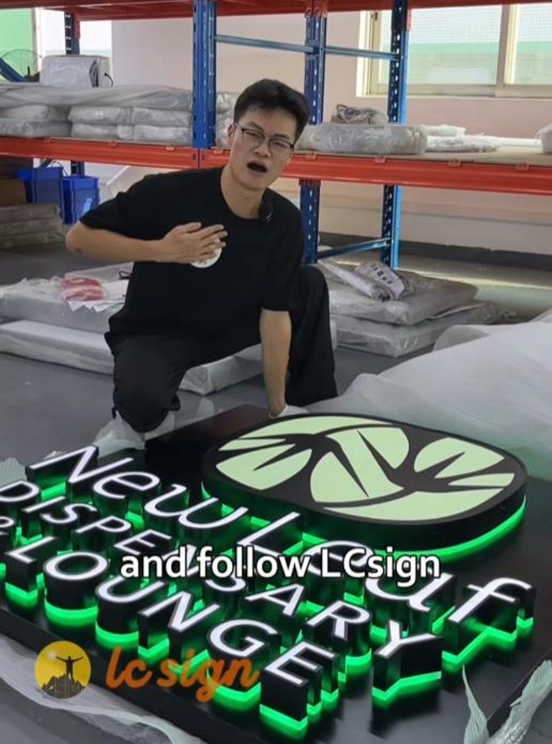 May include: A black rectangular sign featuring the white text New Leaf Dispensary Lounge and a light green leaf logo. The sign has bright green LED backlighting behind the raised letters and logo. The text and follow LCsign is visible.
