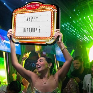 May include: A decorative sign with the text "HAPPY BIRTHDAY" in a marquee style. The sign has a red and gold border with the words "BUFFALO BILLS" at the top. The sign is held up by a person in a sparkly dress.