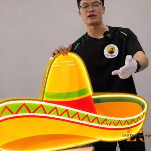 May include: A large, illuminated sombrero sign with a bright yellow brim, a red and green band, and a yellow crown. The sign has the text "Led Custom sign" in the bottom right corner.