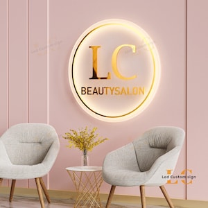 May include: A round, illuminated sign with gold lettering that reads "LC BEAUTYSALON" hangs on a pink wall. Two gray armchairs and a small table with a vase of yellow flowers are in front of the wall.