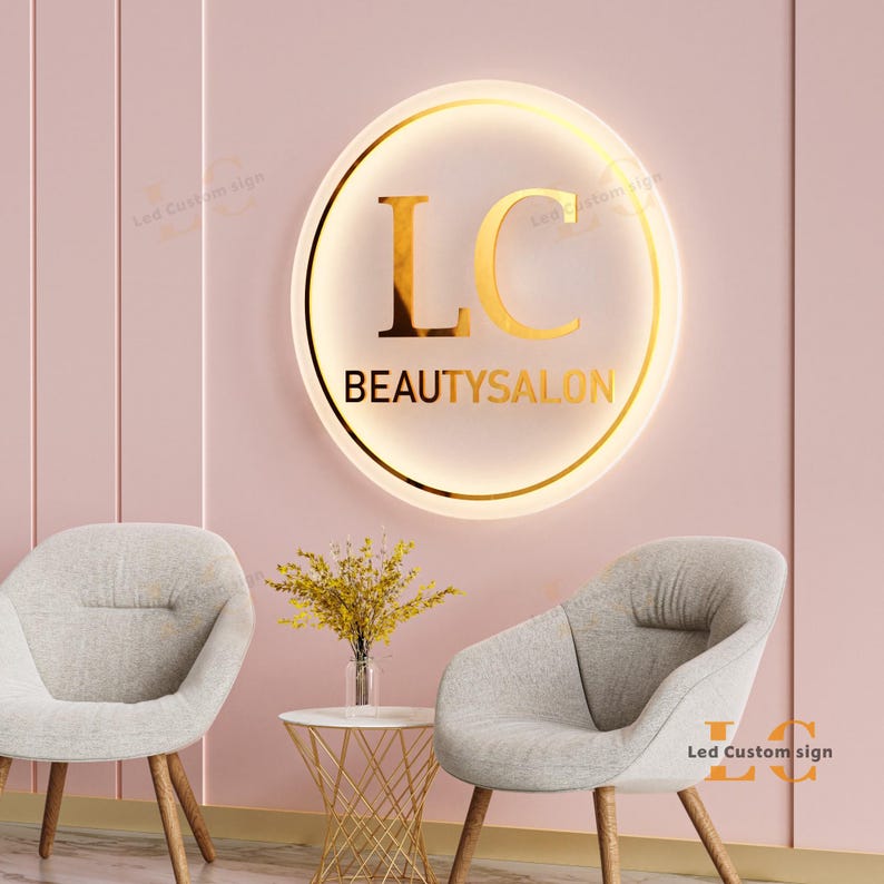 May include: A round, illuminated sign with gold lettering that reads "LC BEAUTYSALON" hangs on a pink wall. Two gray armchairs and a small table with a vase of yellow flowers are in front of the wall.