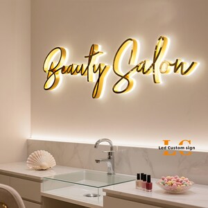 May include: A wall-mounted gold-colored sign with illuminated script lettering that reads Beauty Salon. The sign features a warm glow emanating from behind the letters, which are mounted on a plain wall above a white countertop and sink.