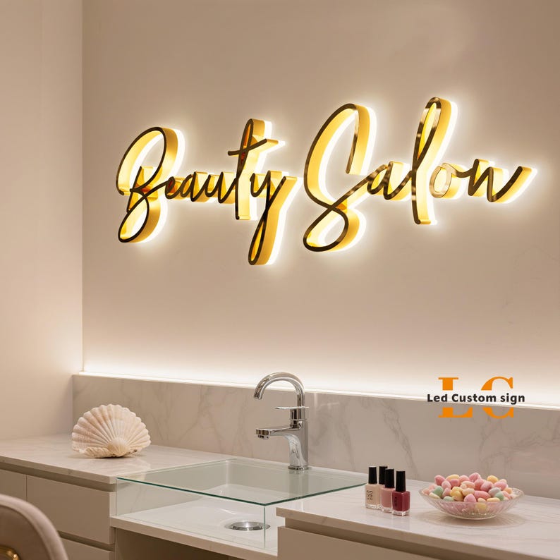 May include: A wall-mounted gold-colored sign with illuminated script lettering that reads Beauty Salon. The sign features a warm glow emanating from behind the letters, which are mounted on a plain wall above a white countertop and sink.