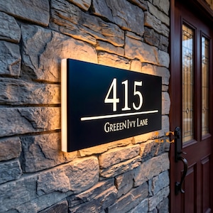May include: A rectangular black address sign with the number "415" in white, illuminated by a warm light. Below the number is the text "GREEN IVY LANE" in white. The sign is mounted on a stone wall.