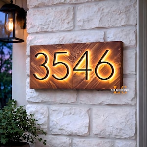 May include: A rectangular wooden house number sign displaying the digits 3546 in a bold, black font. The numbers are backlit with a warm, glowing light that illuminates the wood grain. The text Led Custom sign is visible in the bottom right corner.
