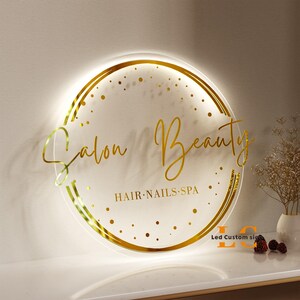 May include: A round, illuminated sign with the words "Salon Beauty" in gold script. The sign also includes the text "HAIR NAILS SPA" and is surrounded by a gold ring and gold dots. The sign is a custom LED sign.