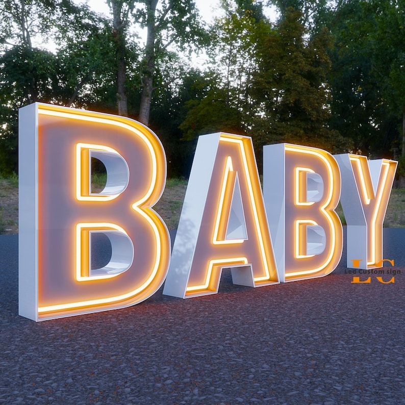 K&ouml;nnte beinhalten: Ein gro&szlig;es, beleuchtetes Schild mit dem Wort &bdquo;BABY&ldquo;. Die Buchstaben sind wei&szlig; mit einem warmen, gelb-orangen Schein, der von innen heraus strahlt. Das Schild steht im Freien auf einer gepflasterten Oberfl&auml;che, mit B&auml;umen im Hintergrund.