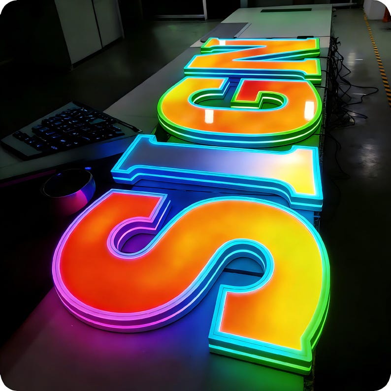 May include: Illuminated "SIGN" letters in a vibrant display. The letters are outlined with neon lights, showcasing a gradient of colors from orange to yellow and green. The letters are on a white surface.