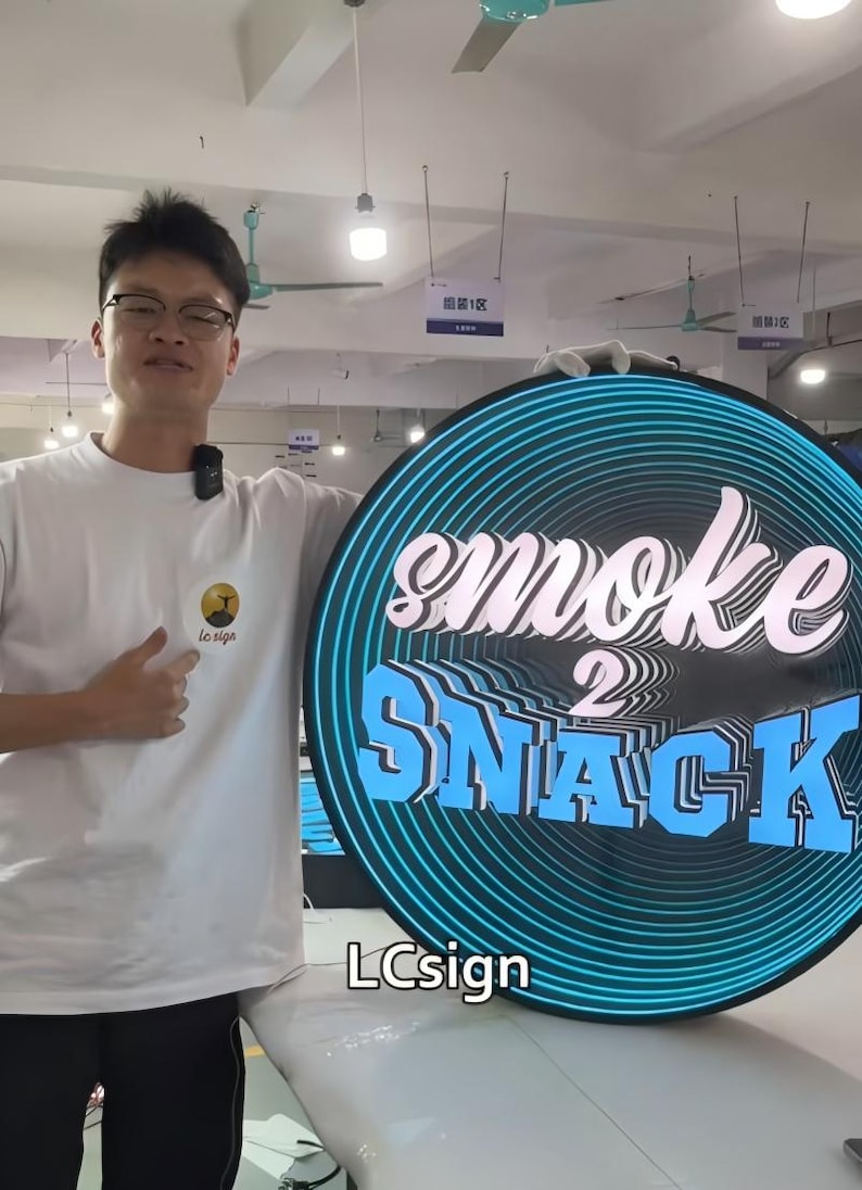May include: A round sign with the words "smoke 2 SNACK" in white and blue, with a blue neon outline. The sign is held by a person wearing a white t-shirt. The sign also has the text "LCsign".