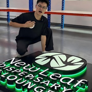 Pu&ograve; includere: Un'insegna luminosa tridimensionale personalizzata con il testo New Leaf Dispensary Lounge in lettere nere in rilievo. L'insegna include un logo a foglia verde brillante e una retroilluminazione a LED verde su una base rettangolare nera.