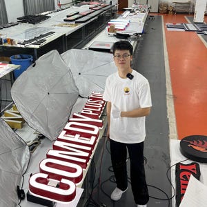 May include: An interior shot of a workshop with a person standing next to a large, illuminated sign that reads "COMEDY PLUS". The sign has a red border and white lettering. Other signs and equipment are visible in the background.