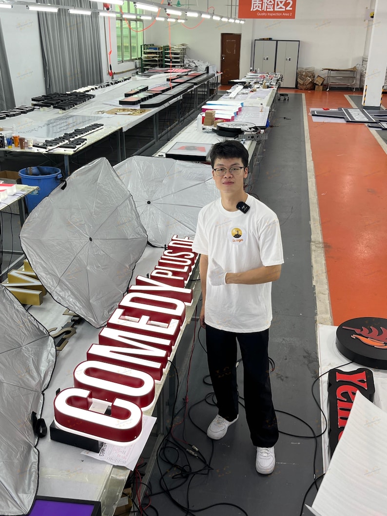 May include: An interior shot of a workshop with a person standing next to a large, illuminated sign that reads "COMEDY PLUS". The sign has a red border and white lettering. Other signs and equipment are visible in the background.